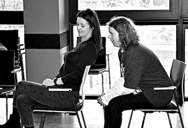 Two workshop participants sitting back-to-back on chairs, leaning into each other's weight in a listening exercise